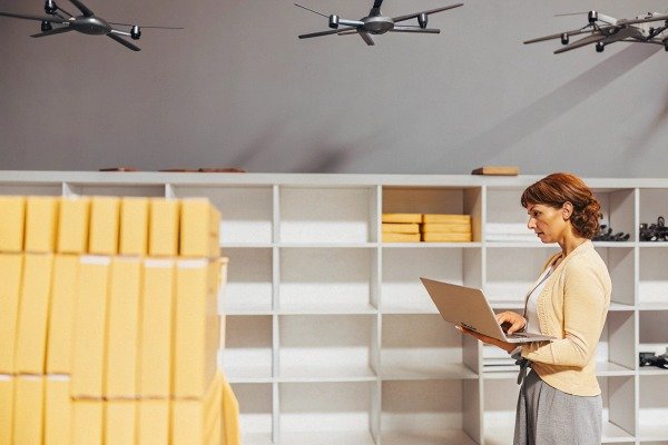 Woman with laptop in a warehouse with drones and stacked boxes (ID#5)