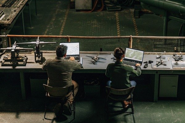 Technicians working on laptops at a long workbench with various drone parts and equipment (ID#3)