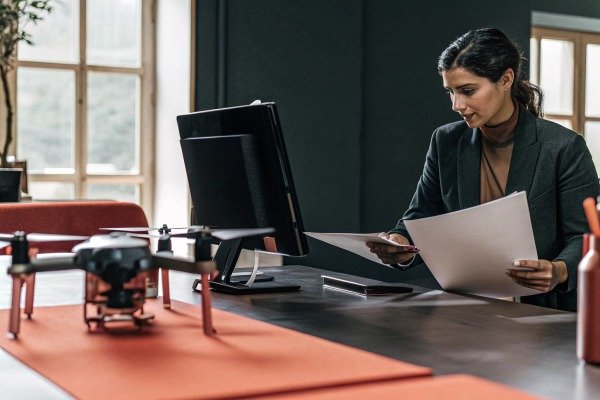 Professional woman reviewing documents at a desk with a modern drone in the foreground (ID#4)