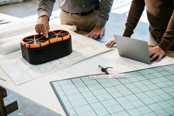 Engineers reviewing drone blueprints and components on a large workspace table (ID#3)
